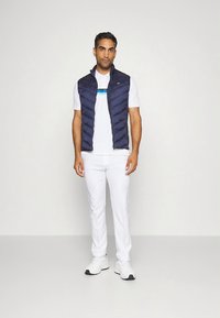 Tamsiai mėlyna paminkšta liemenė ant balto polo marškinėlio su mėlynomis juostelėmis, derinama su baltais kelnėmis ir baltais sportbačiais. Paprastas, prigludęs dizainas.