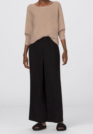 Nainen, jolla on beige pitkähihainen neulepaita, mustat leveälahkeiset housut ja mustat karvaiset avoimet tossut, seisoo yksiväristä taustaa vasten.