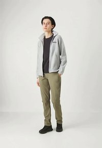 Oseba stoji v svetlo sivi jopici z zadrgo, črni majici, olivno zelenih hlačah in črnih čevljih pred enobarvno svetlo ozadje.