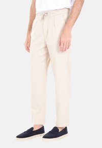 Chino beige con vita regolabile, pieghe sulla parte frontale e tasche laterali. Il tessuto ha una consistenza liscia. Abbinati a scarpe slip-on blu navy.