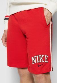 Červené bavlněné šortky s vyšitým logem "NIKE", bočními pruhy a kapsami, hladkou texturou a elastickým pasem.