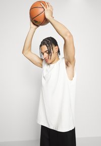 Locker sitzendes, ärmelloses weißes Baumwollshirt mit rundem Halsausschnitt. Die Person hält einen Basketball über ihrem Kopf.