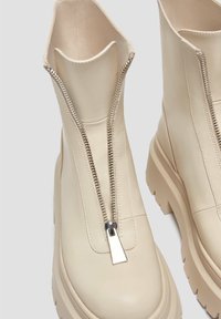 Bottines couleur crème avec semelles épaisses et fermetures éclair métalliques à l'avant partiellement ouvertes, montrant une texture de cuir lisse.