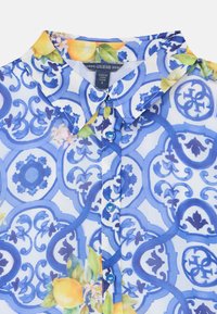 Μπλε μπλούζα με σχέδια που περιλαμβάνουν λουλουδάτες λεπτομέρειες σε κίτρινο και πράσινο, ελαφρύ διαφανές ύφασμα, μυτερό γιακά και μπλε κουμπιά.