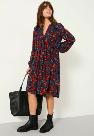 Blumendruckkleid in Marineblau mit roten und orangefarbenen Blumen, plissiertes Design, lange Ärmel, kombiniert mit schwarzen Stiefeletten und einer Tragetasche.