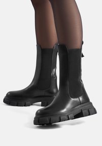 Czarne botki skórzane z elastycznymi panelami bocznymi, grubymi podeszwami i matowym wykończeniem. Surowy design z fakturowanymi detalami.