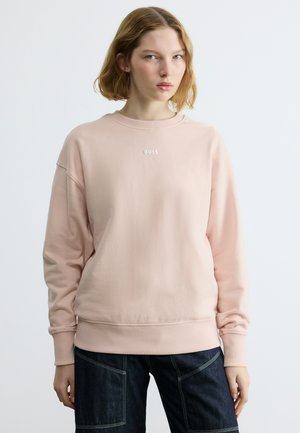 Νεαρή γυναίκα με κοντά μαλλιά που φοράει ένα ανοιχτό ροζ φούτερ BOSS και σκούρο τζιν, στέκεται μπροστά σε ένα απλό λευκό φόντο.