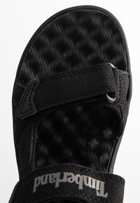 Svart sandal med en strukturerad fotbädd med ett diamantmönster, justerbara remmar och en logotypdetalj på remmen.