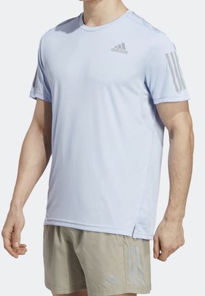 Mann trägt ein hellblaues Adidas kurzärmeliges Sportshirt und beige Shorts mit Adidas-Logos sowie den drei Streifen an Ärmeln und Shorts.