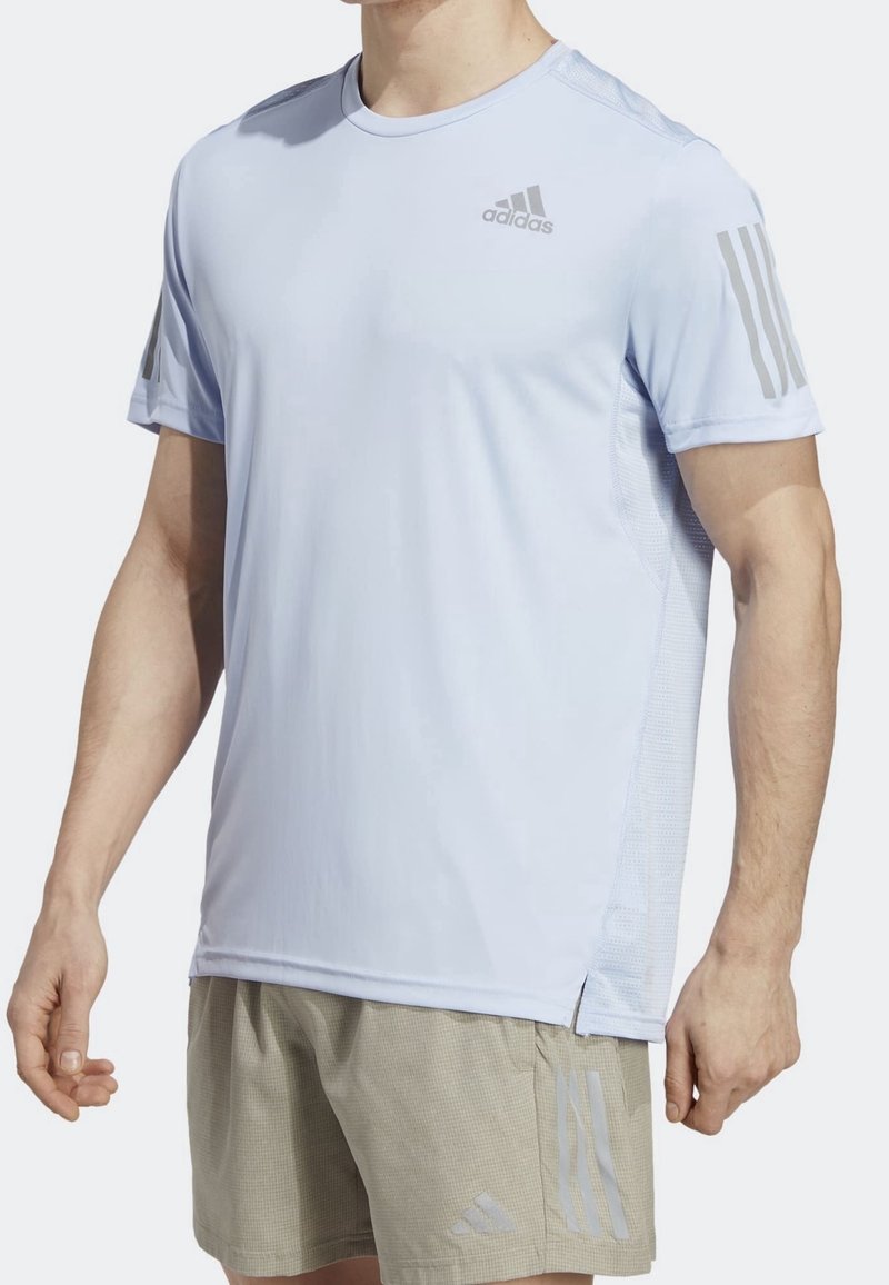 Homme portant un t-shirt de sport Adidas à manches courtes bleu clair et un short beige avec des logos Adidas et trois bandes sur les manches et le short.