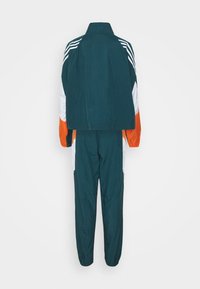 Survêtement vert foncé avec manches blanches et orange, présentant des rayures blanches sur les épaules et des poignets élastiques sur le pantalon et les manches, vu de dos.