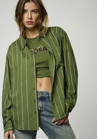 Camicia verde a righe con dettagli bianchi, abbinata a un top verde corto. Indossata con jeans denim dal texture sbiancato.