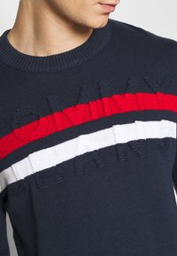 Pull en maille bleu marine avec un logo en relief et des rayures horizontales rouge et blanc sur la poitrine. Col et poignets côtelés.