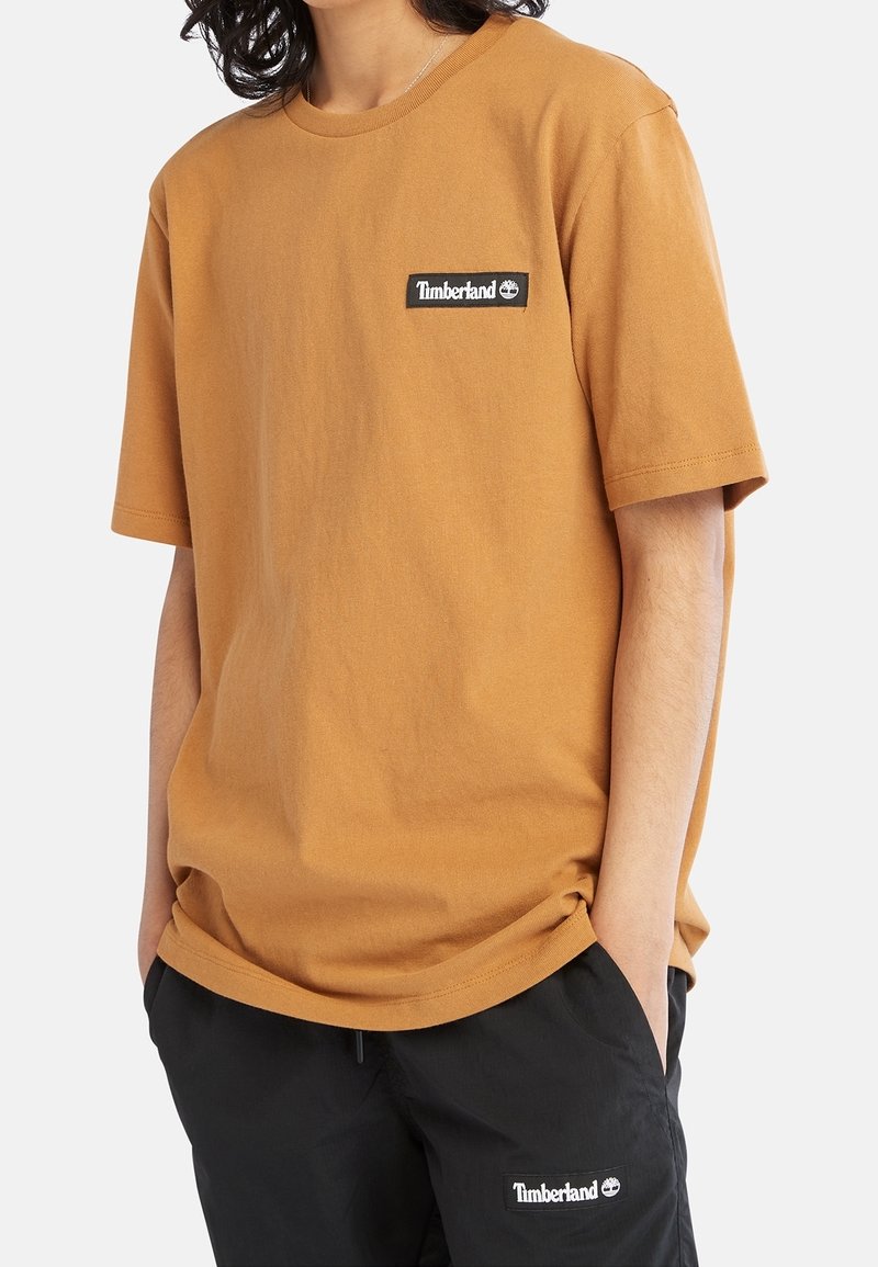 Personne portant un t-shirt Timberland marron et un pantalon Timberland noir, les mains dans les poches, visible du cou jusqu'à mi-cuisse.
