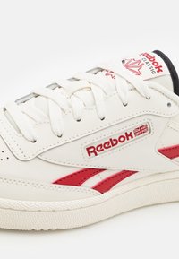White leather sneaker with red accents, textured sole, and Reebok branding. Features round laces and a padded tongue with logo detail.
