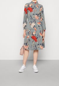 Kobieta ubrana w szarą, kwiatową sukienkę midi z czerwonymi i różowymi kwiatami, białe trampki i trzymająca małą różową torebkę przy boku.