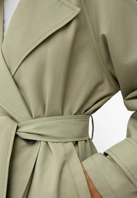 Olivově zelený trenčkot se strukturovaným designem, širokými klopami, opaskem v pase a střiženými rukávy, s hladkou texturou a minimálním kováním.