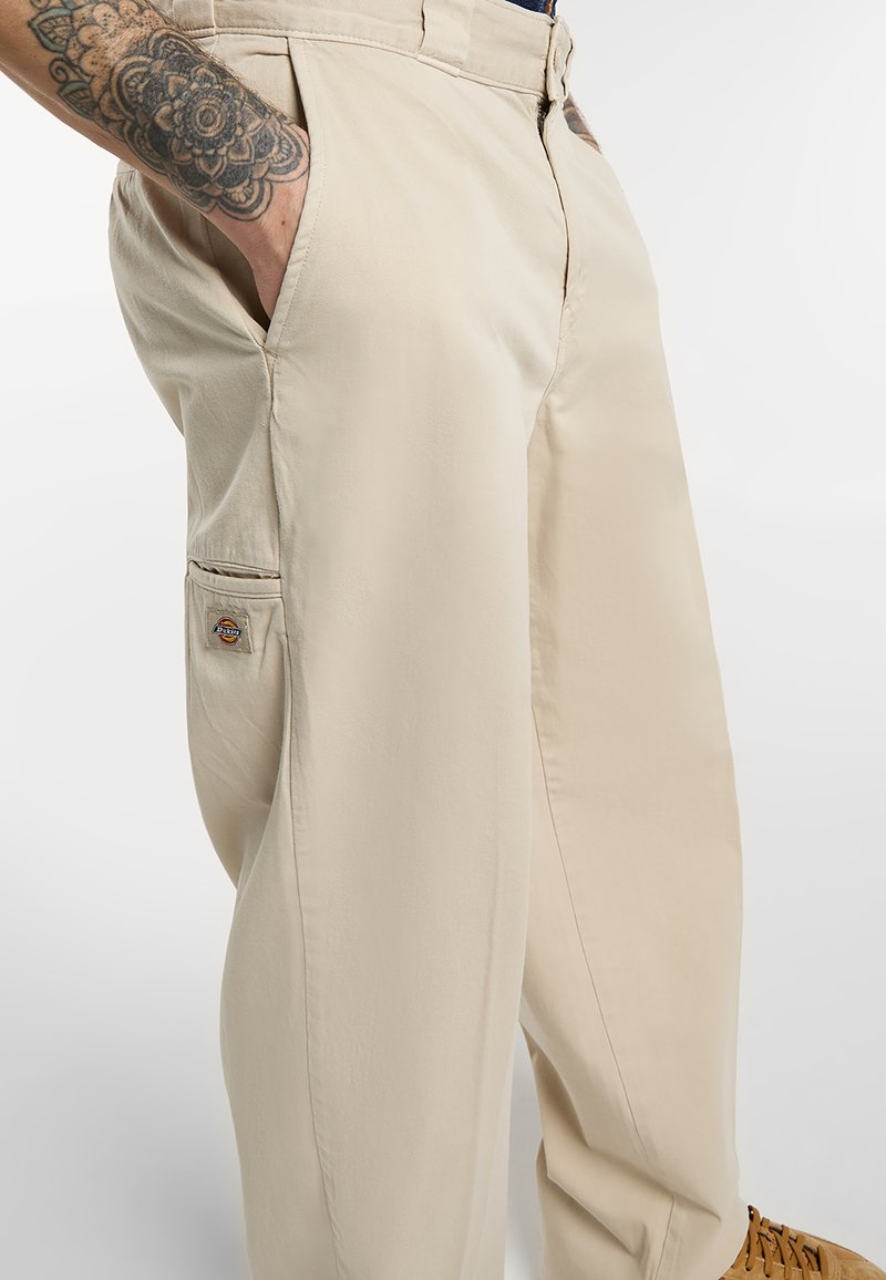 Pantaloni da lavoro beige con tasca laterale e logo, indossati da una persona con avambraccio tatuato e scarpe marroni, in piedi davanti a uno sfondo semplice.