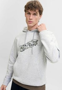 Młody m�ężczyzna z kręconymi włosami, ubrany w jasnoszary hoodie z tekstem w stylu 3D "JACK & JONES 1990", trzymający sznurek od kaptura w okolicach szyi, na neutralnym tle.