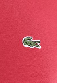 Rode katoenen stof met een gestructureerde afwerking, met een groen geborduurd krokodillenlogo met een witte omlijning.