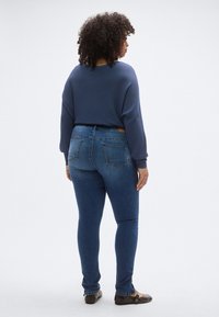 Kék bordázott pulóver ejtett vállakkal, sötétkék szűk farmernadrággal, amelynek hátsó zsebei és barna öve van. Barna lapos cipő.