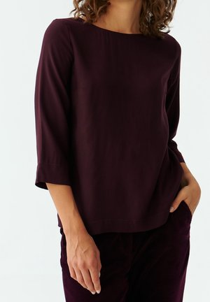 Kobieta ubrana w ciemnofioletową bluzkę z długim rękawem i dekoltem typu łódka oraz pasujące spodnie, stojąca z jedną dłonią w kieszeni, a drugą swobodnie opuszczoną.