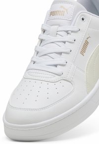Gros plan sur une sneaker Puma blanche avec un bout perforé, une languette rembourrée, des lacets blancs et un logo Puma doré sur la languette et sur le côté.