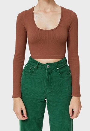 Femme portant un crop top côtelé marron à manches longues et un pantalon en velours côtelé vert taille haute, debout devant un fond uni.