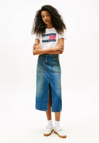 T-shirt blanc avec un design graphique rouge, bleu et blanc, associé à une jupe en denim longueur genou avec une fente à l'avant et des poches. Baskets blanches.