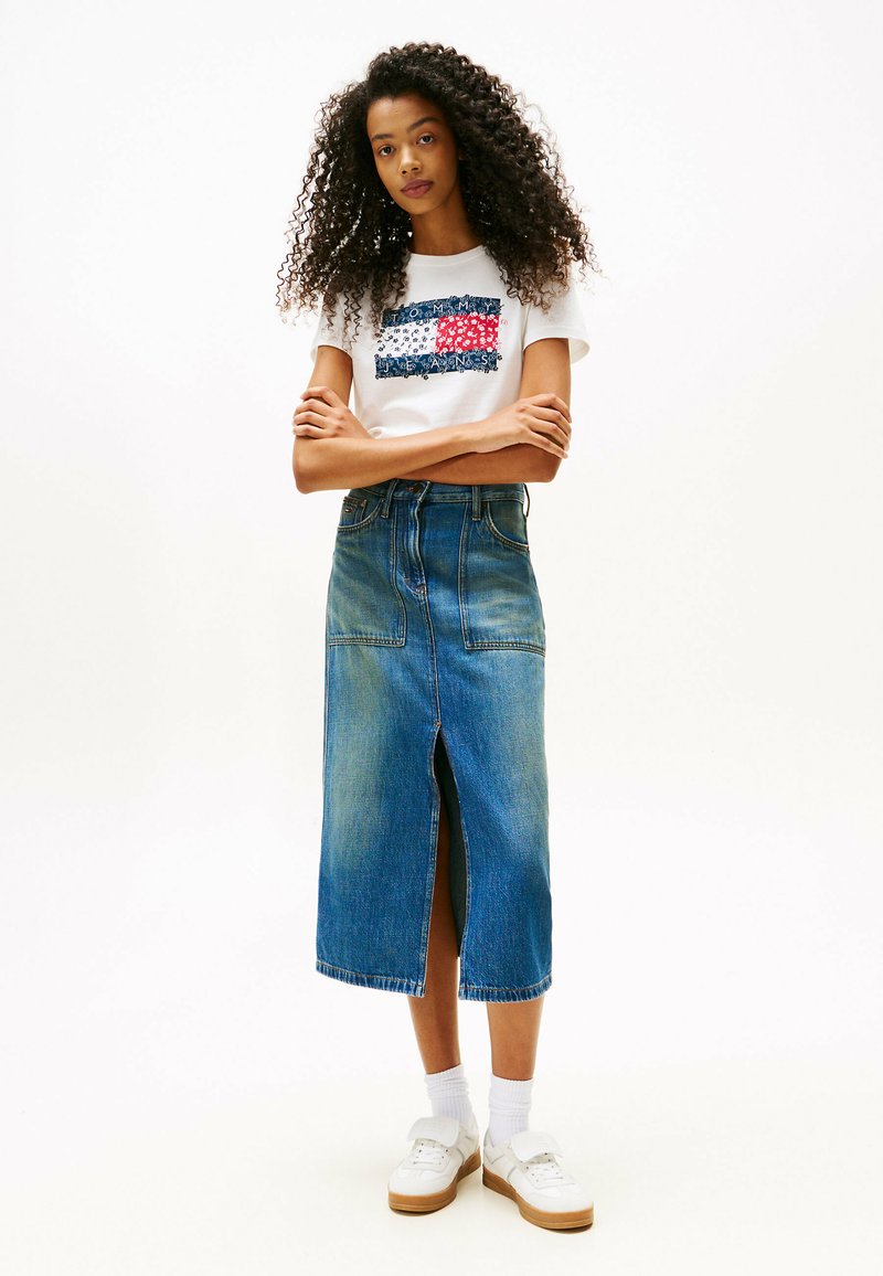 T-shirt blanc avec un design graphique rouge, bleu et blanc, associé à une jupe en denim longueur genou avec une fente à l'avant et des poches. Baskets blanches.