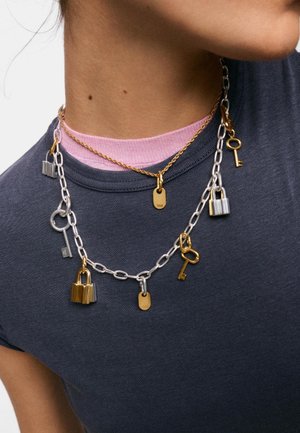 Primer plano de una persona que lleva collares superpuestos con cadenas de plata y oro, candados, llaves y una etiqueta grabada con "TOUS."