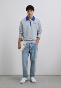 Jonge man in een grijze sweatshirt met blauwe kraag, lichtblauwe jeans, witte sneakers, die in één hand een notitieboek en tijdschriften vasthoudt.