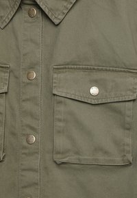 Camicia verde oliva realizzata in cotone, con colletto a punta, due tasche a pattina e chiusura con bottoni color oro.