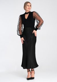 Schwarzes Kleid mit durchsichtigen, gepunkteten Puffärmeln, einem V-Ausschnitt und einem figurschmeichelnden, glatten Satinrock. Getragen mit schwarzen Riemchen-Heels.