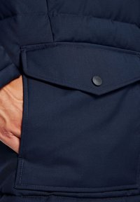 Granatowa pikowana kurtka z widoczną kieszenią z przodu. Kieszeń ma klapkę i czarny guzik do bezpiecznego zapięcia.