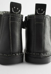 Bottes en cuir noir avec une finition texturée, des coutures blanches et un logo de smiley sur la tirette à l'arrière. Semelles en caoutchouc plates.