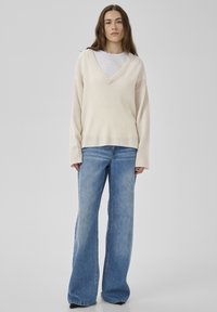 Pull crème à col en V surdimensionné avec ourlet côtelé, associé à un jean bleu clair évasé. Les deux vêtements présentent une coupe décontractée et une texture douce.