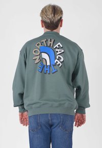Felpa verde con una grande grafica colorata "The North Face" sulla schiena. Presenta polsini e orlo a costine, indossata con jeans blu.