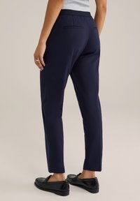 Persona che indossa pantaloni dritti blu navy, una maglietta grigia e mocassini neri, in piedi con la schiena rivolta verso la telecamera su uno sfondo neutro.