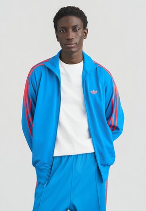 Sininen vetoketjullinen verkkaritakki punaisilla raidoilla, jossa on kirjailtu logo rinnassa. Yhdistetty samansävyisiin sinisiin shortseihin.