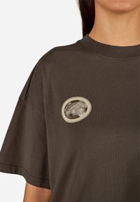 Brązowy bawełniany t-shirt z krótkim rękawem, z okrągłym haftowanym naszywką przedstawiającą wzór domu w beżowym i szarym kolorze na klatce piersiowej.