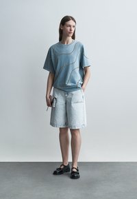 Kvinde iført en blå oversized t-shirt med kædemønstre, lyse denim Bermuda-shorts, sorte sko, og holder en lille grå clutch.