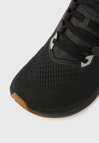 Μαύρο αθλητικό παπούτσι με πλεκτό πλέγμα στο πάνω μέρος, μαύρα κορδόνια, ανακλαστικές λεπτομέρειες κοντά στα κορδόνια και καφέ λαστιχένιο προστατευτικό άκρου στην μύτη, σε λευκό φόντο.