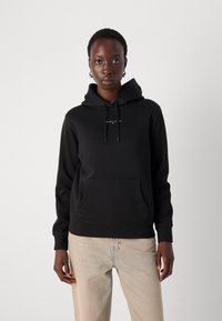 Sudadera negra con capucha, bolsillo frontal y cordón. Presenta un pequeño logo blanco en el pecho. Combinada con unos vaqueros de color claro.