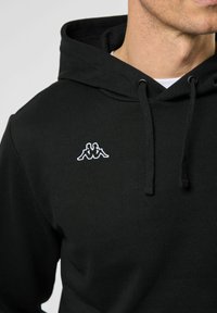 Zwarte hoodie van katoen, met een trekkoordcapuchon en een wit geborduurd logo op de borst. Soepele textuur en moderne pasvorm.