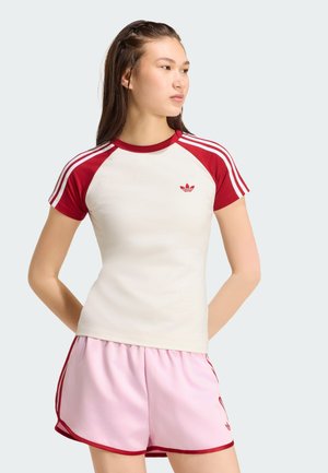 Ung kvinde står med hænderne bag ryggen iført hvid og rød Adidas T-shirt og lyserøde shorts med røde kanter.