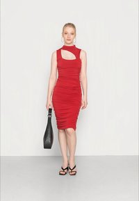 Vestido ajustado vermelho com drapeado, design sem mangas e detalhe recortado na gola. Usado com sandálias pretas com tiras e segurando uma bolsa preta.
