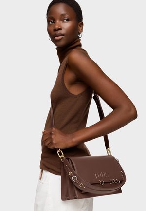 Mujer con suéter sin mangas de cuello alto marrón y pantalones blancos, llevando un bolso de cuero marrón de TOUS en el hombro.