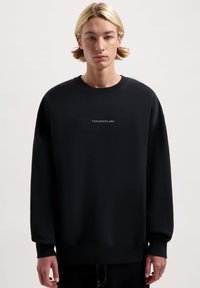 Zwarte sweatshirt gemaakt van een zachte stof, met een ronde halslijn en lange mouwen, waarop "TOMORROWLAND" in het wit op de voorkant is gedrukt.