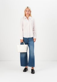 Camicia bianca a maniche lunghe, jeans blu a gamba larga, mocassini neri, tiene in mano una piccola borsa bianca. Camicia rosa chiaro con dettagli di bottoni.